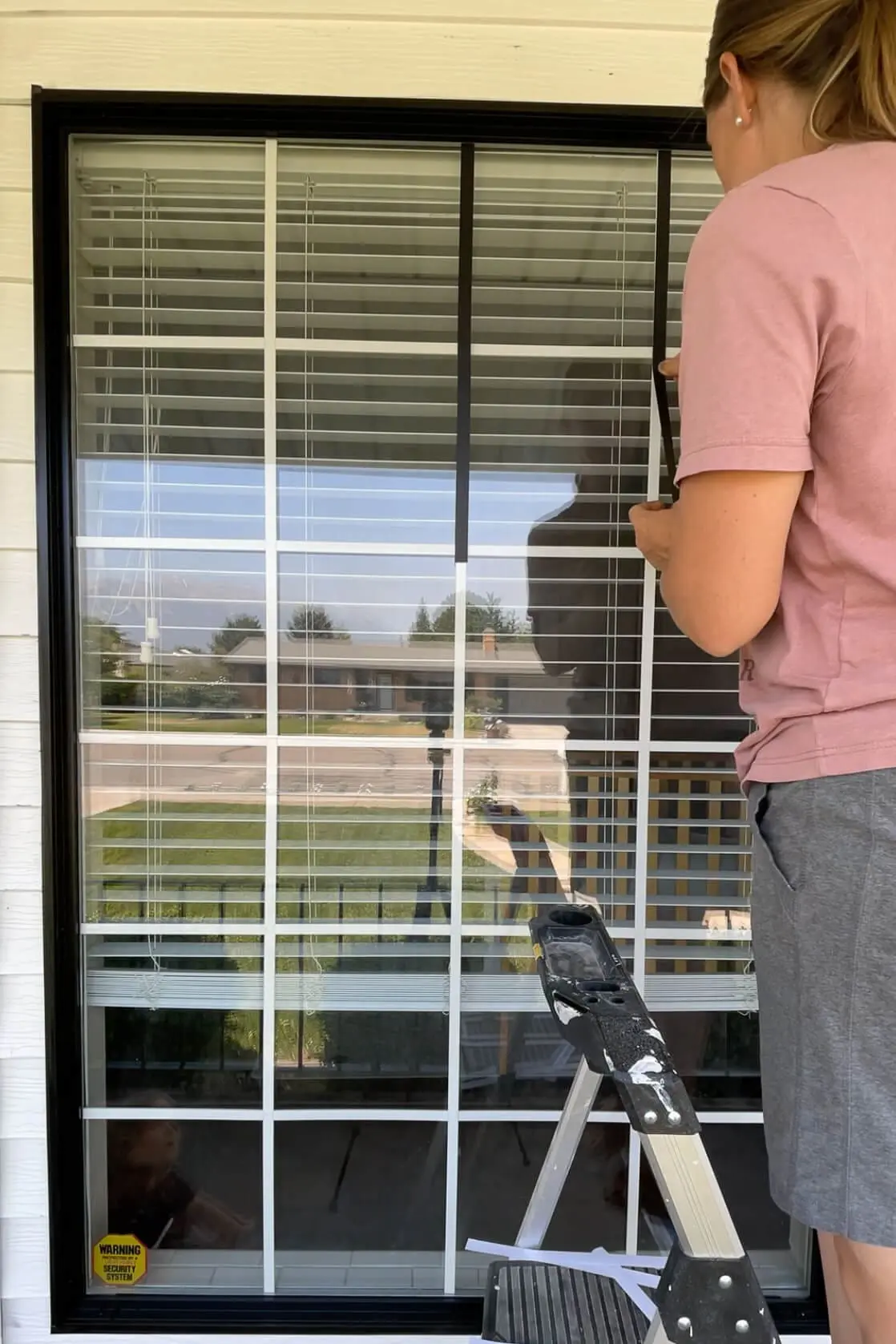 Woman applying a black vinyl strip vertically over a white window grid using a ladder during a DIY window upgrade.